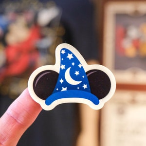 Może przedstawiać: Naklejka z niebieskim i białym kapeluszem Myszki Miki z półksiężycem i gwiazdami. Naklejkę trzyma palec.