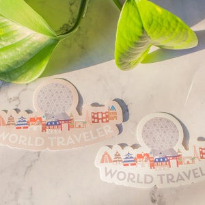 Op de afbeelding: Twee witte stickers met een kleurrijke illustratie van een stadsbeeld en de tekst "WORLD TRAVELER". De stickers tonen een gestileerde versie van de Spaceship Earth attractie in Epcot.