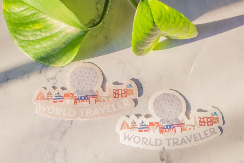 Op de afbeelding: Twee witte stickers met een kleurrijke illustratie van een stadsbeeld en de tekst "WORLD TRAVELER". De stickers tonen een gestileerde versie van de Spaceship Earth attractie in Epcot.