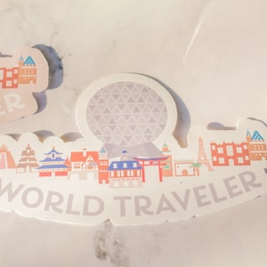 Op de afbeelding: Een witte sticker met een kleurrijke illustratie van verschillende gebouwen en de tekst "WORLD TRAVELER". De sticker heeft een grijze en witte gepatenteerde bol in het midden.