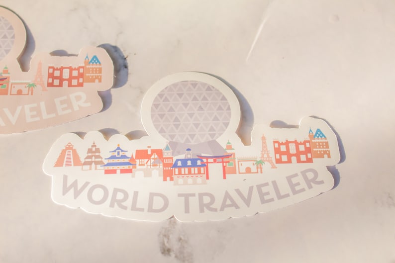 Op de afbeelding: Een witte sticker met een kleurrijke illustratie van verschillende gebouwen en de tekst "WORLD TRAVELER". De sticker heeft een grijze en witte gepatenteerde bol in het midden.