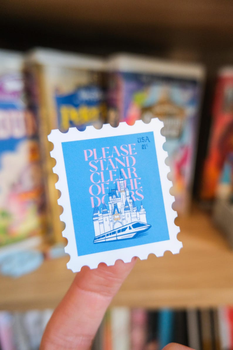 Pode incluir: Um selo postal quadrado com fundo azul e o texto "PLEASE STAND CLEAR OF THE DOORS". O selo apresenta uma ilustra&ccedil;&atilde;o de um castelo e um monotrilho. O selo est&aacute; marcado "USA 21".