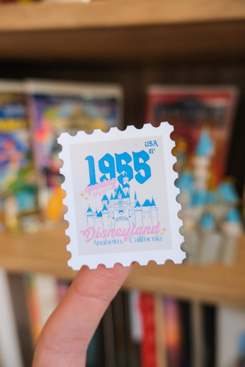 Pode incluir: Um selo postal branco com um castelo azul, o texto "1955" e "Disneyland Anaheim, Calif&oacute;rnia". O selo tamb&eacute;m inclui o texto "Abertura 17 de julho" e "USA 21&cent;".