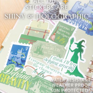 Op de afbeelding: Een verzameling glanzende en holografische stickers met Wizard of Oz-thema's. De stickers bevatten afbeeldingen van een heks, een bezem, koffers en de Emerald City. Tekst bevat "Defying Gravity" en "Everyone deserves the chance to fly."