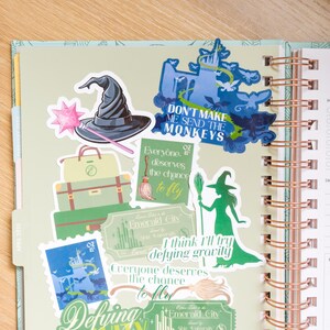 Op de afbeelding: Een planner open op een pagina met kleurrijke stickers. Stickers zijn onder meer een heksenhoed, een toverstaf, bagage en zinnen als "Don't Make Me Send The Monkeys" en "Defying Gravity". De planner heeft een spiraalbinding.