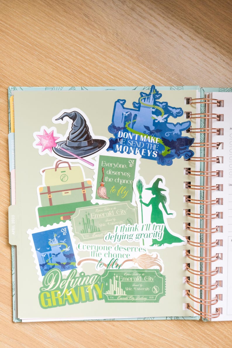 Op de afbeelding: Een planner open op een pagina met kleurrijke stickers. Stickers zijn onder meer een heksenhoed, een toverstaf, bagage en zinnen als "Don't Make Me Send The Monkeys" en "Defying Gravity". De planner heeft een spiraalbinding.