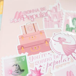 Op de afbeelding: Roze stickers met de tekst "Gonna Be Popular" en andere zinnen. De stickers tonen afbeeldingen van koffers, een kasteel, een heksenhoed, een toverstaf en een silhouet van een vrouw. Op een roze achtergrond.