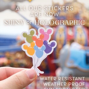 Op de afbeelding: Een close-up van een hand die een sticker vasthoudt met kleurrijke Mickey Mouse ballonnen. De sticker is glanzend en holografisch. De tekst op de sticker luidt "NEW! ALL OUR STICKERS ARE NOW SHINY or HOLOGRAPHIC WATER RESISTANT WEATHER PROOF SUN PROTECTED GREAT FOR PLANNERS, LAPTOPS & WATER BOTTLES!"