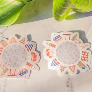 Op de afbeelding: Twee witte stickers met een cirkelvormig ontwerp met een grijze geodetische koepel omringd door kleurrijke illustraties van beroemde bezienswaardigheden van over de hele wereld.