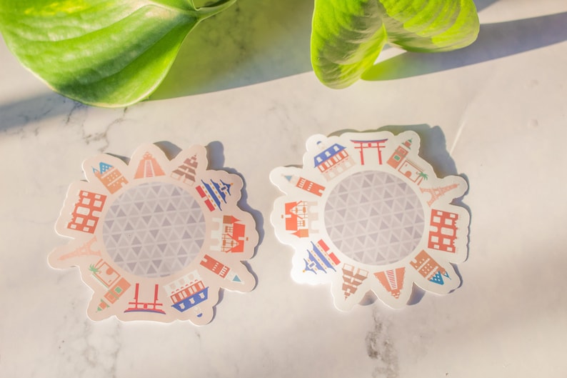 Op de afbeelding: Twee witte stickers met een cirkelvormig ontwerp met een grijze geodetische koepel omringd door kleurrijke illustraties van beroemde bezienswaardigheden van over de hele wereld.