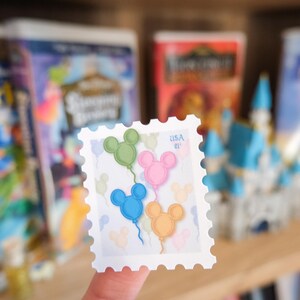 Pode incluir: Um selo postal branco com quatro bal&otilde;es em forma de Mickey Mouse em verde, rosa, azul e laranja. O selo tem o texto "USA 21" impresso. O selo &eacute; segurado em frente a um fundo desfocado.
