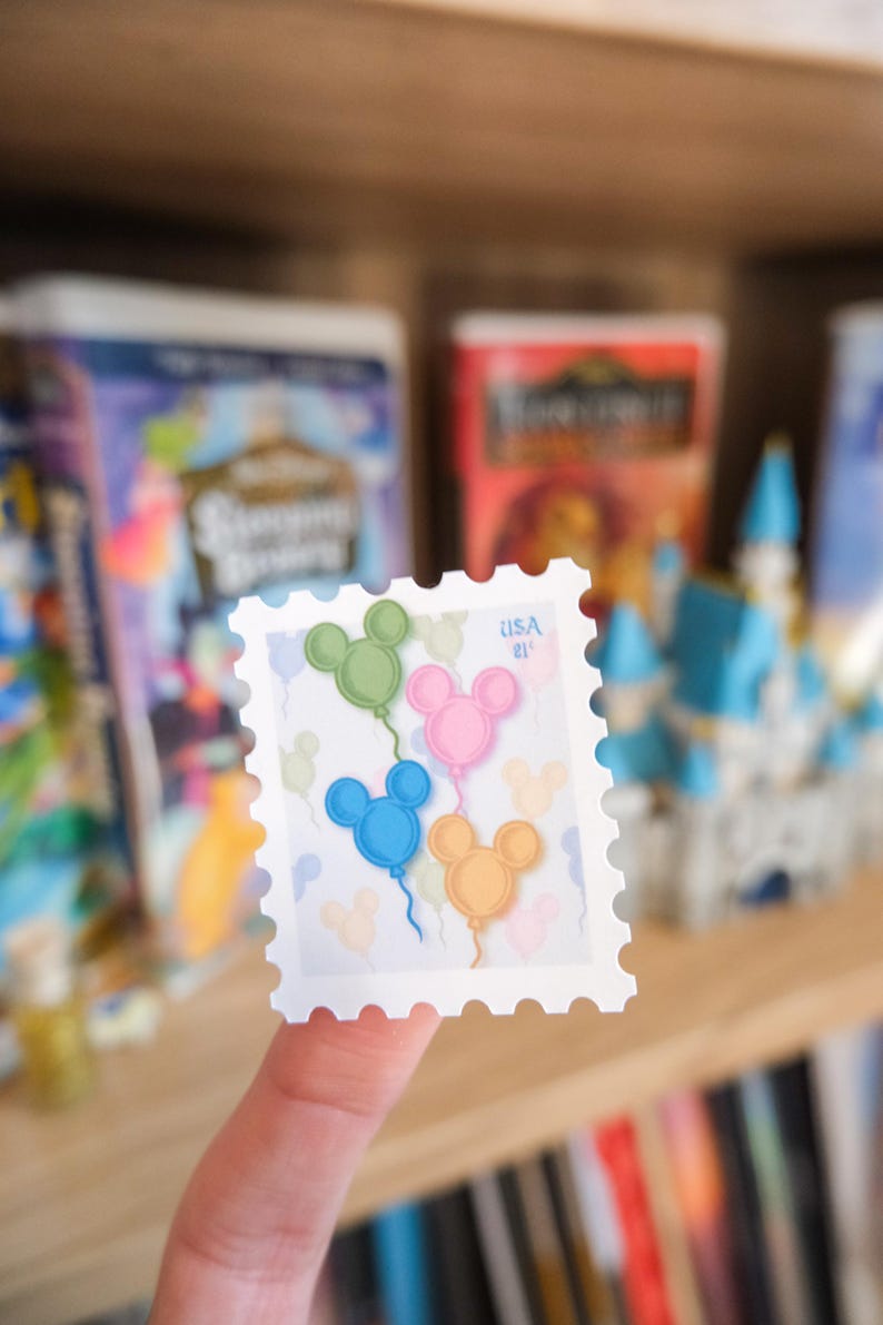 Pode incluir: Um selo postal branco com quatro bal&otilde;es em forma de Mickey Mouse em verde, rosa, azul e laranja. O selo tem o texto "USA 21" impresso. O selo &eacute; segurado em frente a um fundo desfocado.
