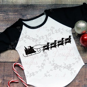 May include: A white and black raglan-style shirt with a Christmas theme. The shirt features a Santa Claus in a sleigh pulled by dachshunds. The sleeves are black with ruffled cuffs. Two Christmas ornaments and a candy cane are also present.