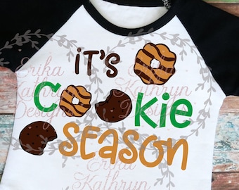 May include: White long-sleeved shirt with black sleeves. The shirt has a graphic design of cookies and the text "It's Cookie Season".