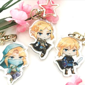 May include: Three cartoon character keychains featuring a blonde haired character in three different outfits. The character is wearing a blue and gold outfit, a blue and brown outfit, and a blue and black outfit. Each keychain has a gold star charm attached.