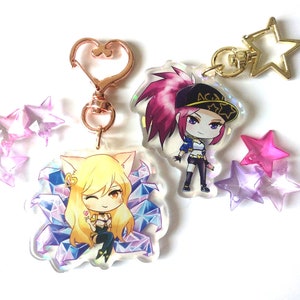 May include: Two colorful acrylic keychains featuring anime characters from the video game League of Legends. The keychain on the left features a character with yellow hair and cat ears, surrounded by blue and purple crystals. The keychain on the right features a character with pink hair wearing a black and white baseball cap with the word "ACE" on it. Both keychains are attached to gold-colored star-shaped charms.