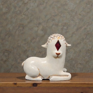 May include: A ceramic lamb figurine in a reclining pose. The lamb is primarily white with brown accents and a red diamond-shaped detail where an eye would be. The lamb has a textured head and is resting on a wooden surface.
