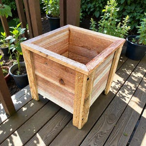 May include: A square wooden planter box with a natural wood finish. The planter has a rectangular shape with four legs and is constructed from horizontal planks. The box is empty and ready for planting. The planter is sitting on a wooden deck.