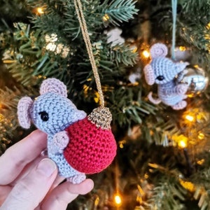 Puede incluir: Un adorno de ratón gris de crochet que sostiene un adorno rojo de crochet. El adorno cuelga de una cuerda dorada en un árbol de Navidad.