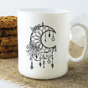 May include: White ceramic mug with a black and white illustration of a crescent moon with a dreamcatcher design. The moon is decorated with stars and feathers.