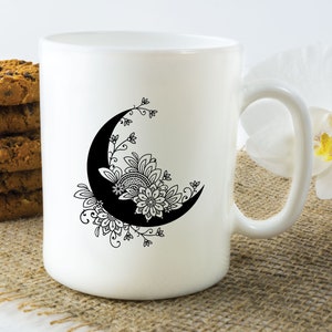 May include: White ceramic mug with a black and white design of a crescent moon with floral details. The mug is sitting on a brown burlap surface.