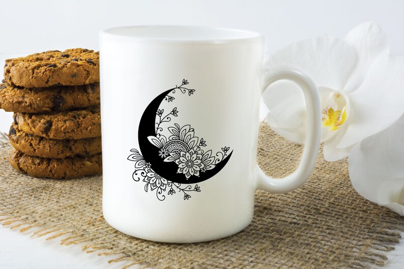 May include: White ceramic mug with a black and white design of a crescent moon with floral details. The mug is sitting on a brown burlap surface.