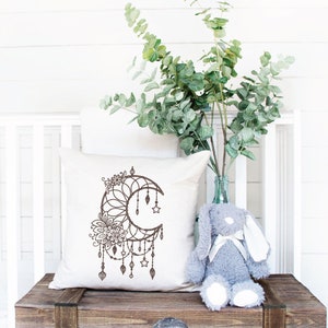 May include: A white throw pillow with a brown dreamcatcher design featuring a crescent moon, stars, and feathers. The pillow is sitting on a wooden bench with a gray stuffed bunny rabbit.