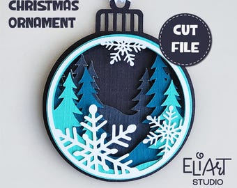 May include: Layered Christmas ornament cut file design featuring a blue and white snowflake design with a forest scene inside. The design is for a layered paper craft project.  SVG, DXF, and PNG file formats are included.  EliArt Studio.