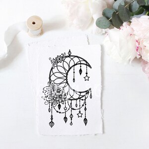 May include: A black and white illustration of a crescent moon with intricate floral details and dangling stars and beads. The illustration is on a white background.