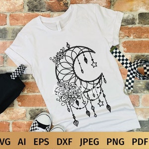 May include: A white t-shirt with a black and white graphic of a crescent moon with floral details and dangling beads. The graphic is reminiscent of a dreamcatcher.