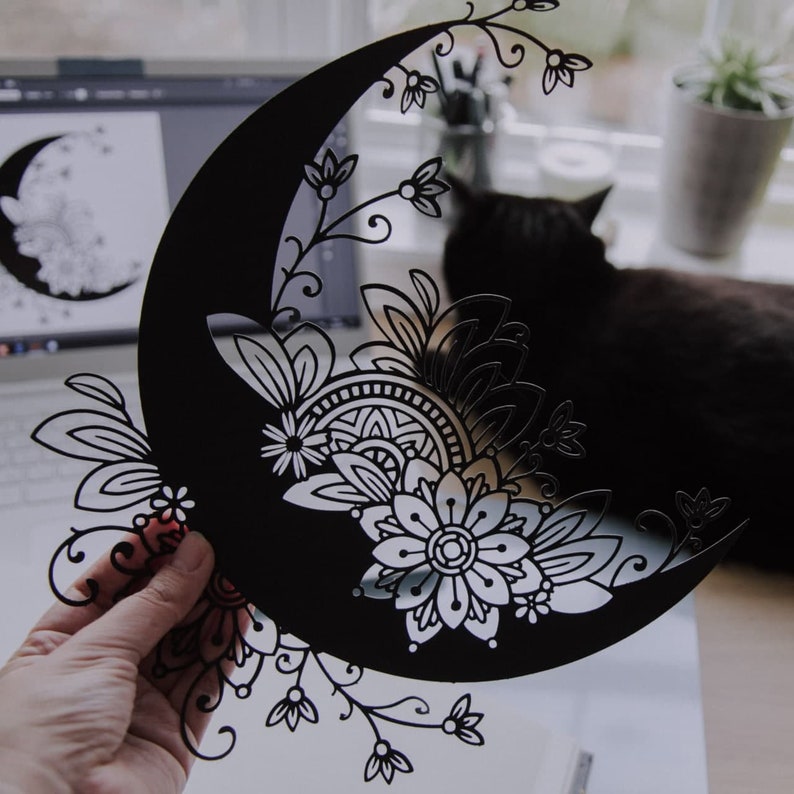 May include: Black paper cut-out of a crescent moon with intricate floral designs. The moon is decorated with a mandala pattern and delicate swirls.