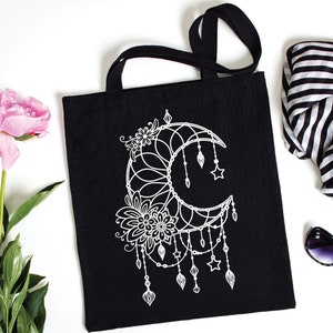 May include: Black canvas tote bag with a white design of a crescent moon with floral details and stars. The bag has two black straps.