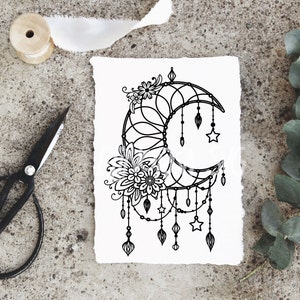 May include: Black and white line art illustration of a crescent moon with floral details and dangling charms. The moon is surrounded by a delicate pattern and has stars and teardrop-shaped charms hanging from it.