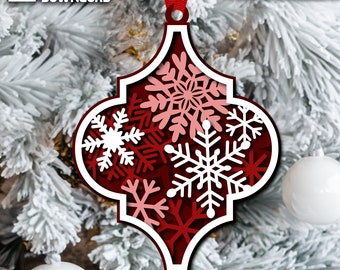 May include: A red and white layered Christmas ornament with snowflake cutouts, hanging from a red ribbon. The ornament has the words "INSTANT DOWNLOAD" and "CUT FILE" on it. The ornament is in front of a snowy Christmas tree.