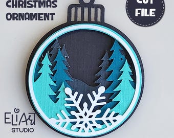 May include: Layered Christmas ornament with a winter forest scene. The ornament features a snowflake and pine trees in shades of blue and white, with a black background. The text "LAYERED CHRISTMAS ORNAMENT" is at the top.
