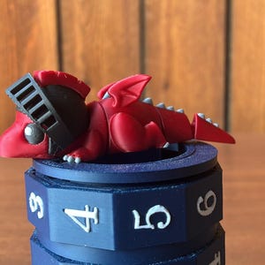 May include: A small red dragon figurine wearing a black knight helmet, resting on top of a dark blue cylindrical puzzle lock. The lock features white numbers 3, 4, 5, and 6 printed on its rotating segments, with a visible gear-like structure.