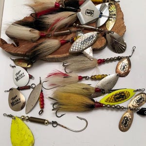 May include: A collection of fishing lures with various designs and colors. The lures feature metallic blades, feathered tails, and hooks. Some lures have the text "Mapps Musky Marabou" on them.