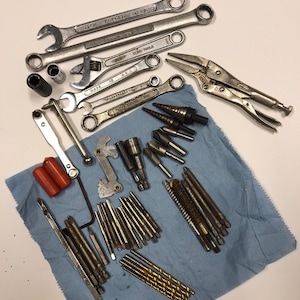 May include: A collection of various metal tools, including wrenches, sockets, pliers, and drill bits, arranged on a blue cloth. The tools are in various sizes and shapes, with some showing signs of use. The image is well-lit, highlighting the details of each tool.
