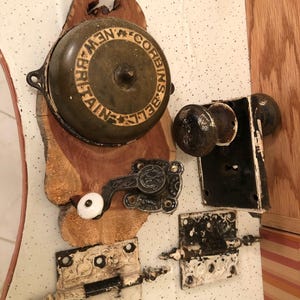May include: An assortment of antique hardware, including a bell with "NEW BRITAIN BELL" text, door knobs, hinges, and a latch. The hardware is mounted on a piece of wood with a natural edge, showcasing a rustic aesthetic.