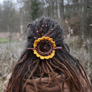 Puede incluir: Una pinza para el pelo de girasol de ganchillo con cuentas de madera marr&oacute;n y un palo de madera. La pinza est&aacute; en las rastas marrones de una persona.