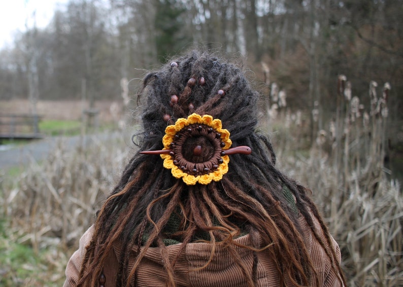 Puede incluir: Una pinza para el pelo de girasol de ganchillo con cuentas de madera marr&oacute;n y un palo de madera. La pinza est&aacute; en las rastas marrones de una persona.