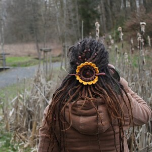 Puede incluir: Una persona con largas rastas marrones lleva una chaqueta marr&oacute;n. Las rastas est&aacute;n adornadas con una pieza de pelo de girasol de ganchillo.