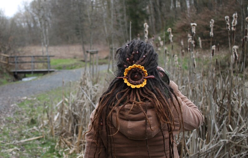 Puede incluir: Una persona con largas rastas marrones lleva una chaqueta marr&oacute;n. Las rastas est&aacute;n adornadas con una pieza de pelo de girasol de ganchillo.