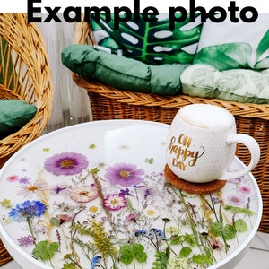 Puede incluir: Mesa auxiliar redonda blanca con una tapa de resina epoxi transparente con flores y vegetaci&oacute;n prensadas. La mesa tiene una base de madera y se encuentra sobre un suelo de baldosas. Una taza blanca con el texto "Oh Happy Day" est&aacute; sobre un posavasos en la mesa.
