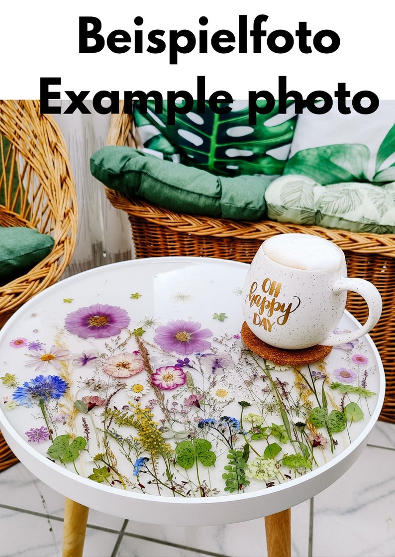 Puede incluir: Mesa auxiliar redonda blanca con una tapa de resina epoxi transparente con flores y vegetaci&oacute;n prensadas. La mesa tiene una base de madera y se encuentra sobre un suelo de baldosas. Una taza blanca con el texto "Oh Happy Day" est&aacute; sobre un posavasos en la mesa.