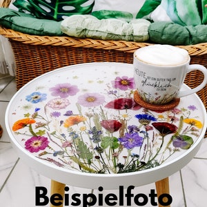 Puede incluir: Mesa auxiliar redonda blanca con una tapa de resina epoxi transparente adornada con flores prensadas en tonos rosa, morado, amarillo y naranja. La mesa tiene patas de madera y se encuentra sobre un suelo de baldosas. El texto en la taza dice "Heute ist ein guter Tag um gl&uuml;cklich zu sein".