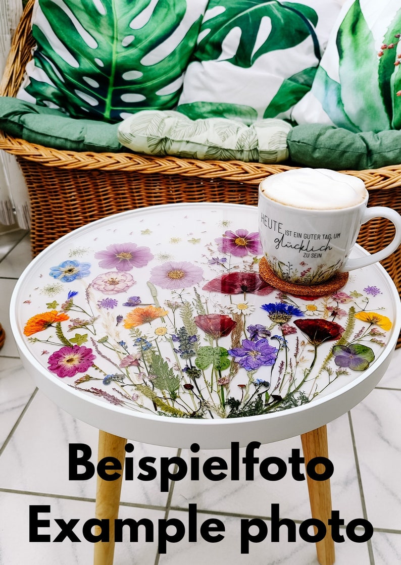 Puede incluir: Mesa auxiliar redonda blanca con una tapa de resina epoxi transparente adornada con flores prensadas en tonos rosa, morado, amarillo y naranja. La mesa tiene patas de madera y se encuentra sobre un suelo de baldosas. El texto en la taza dice "Heute ist ein guter Tag um gl&uuml;cklich zu sein".