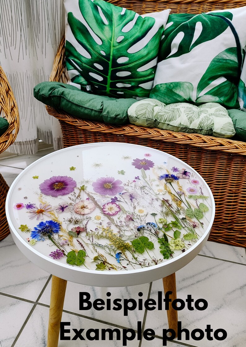 Puede incluir: Mesa auxiliar redonda blanca con patas de madera. La tapa de la mesa est&aacute; hecha de resina transparente con flores y hojas prensadas. El texto "Beispielfoto Example photo" est&aacute; impreso debajo de la mesa.