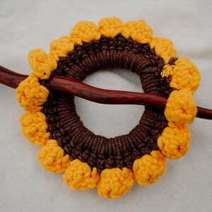 Puede incluir: Un palo de pelo de madera marr&oacute;n con un dise&ntilde;o de girasol tejido a crochet. El girasol est&aacute; hecho de hilo marr&oacute;n y amarillo y tiene un centro elevado.