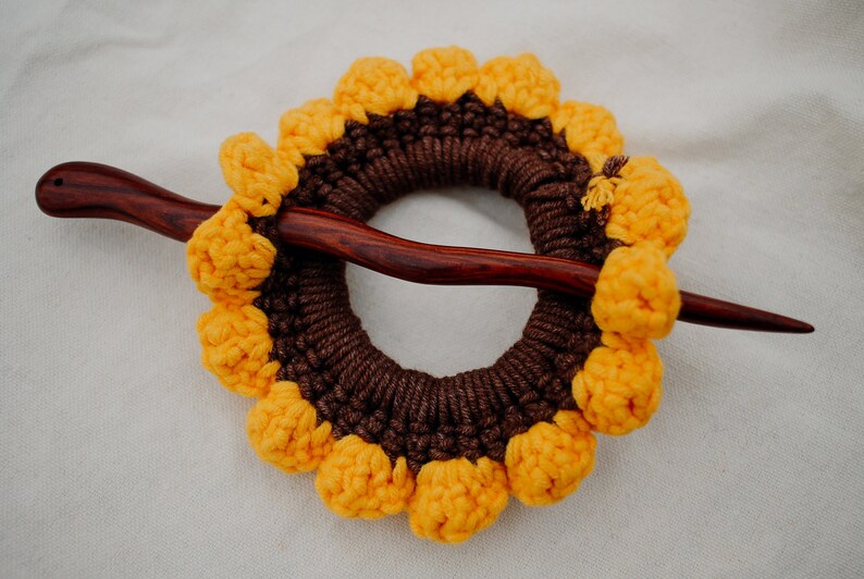 Puede incluir: Un palo de pelo de madera marr&oacute;n con un dise&ntilde;o de girasol tejido a crochet. El girasol est&aacute; hecho de hilo marr&oacute;n y amarillo y tiene un centro elevado.
